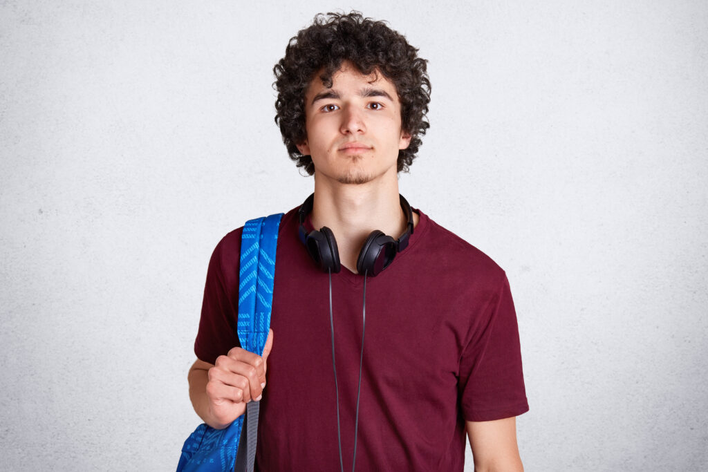 Close up portrait of young guy with curly hair, wearing headphones, maroon t shirt, holds blue racksack, travels around country, stands isolated on white wall, looks directly at camera. Turism concept